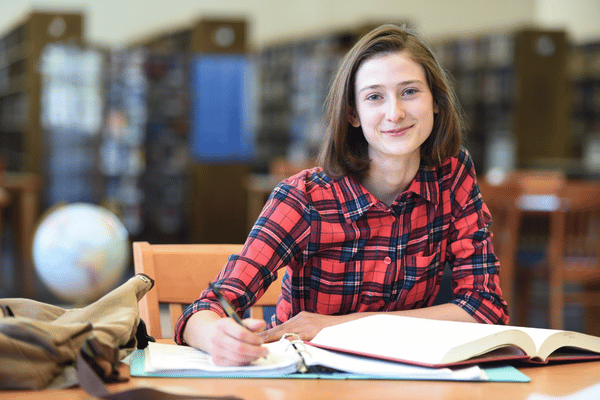 Student studying at a library table with open books and notebooks.